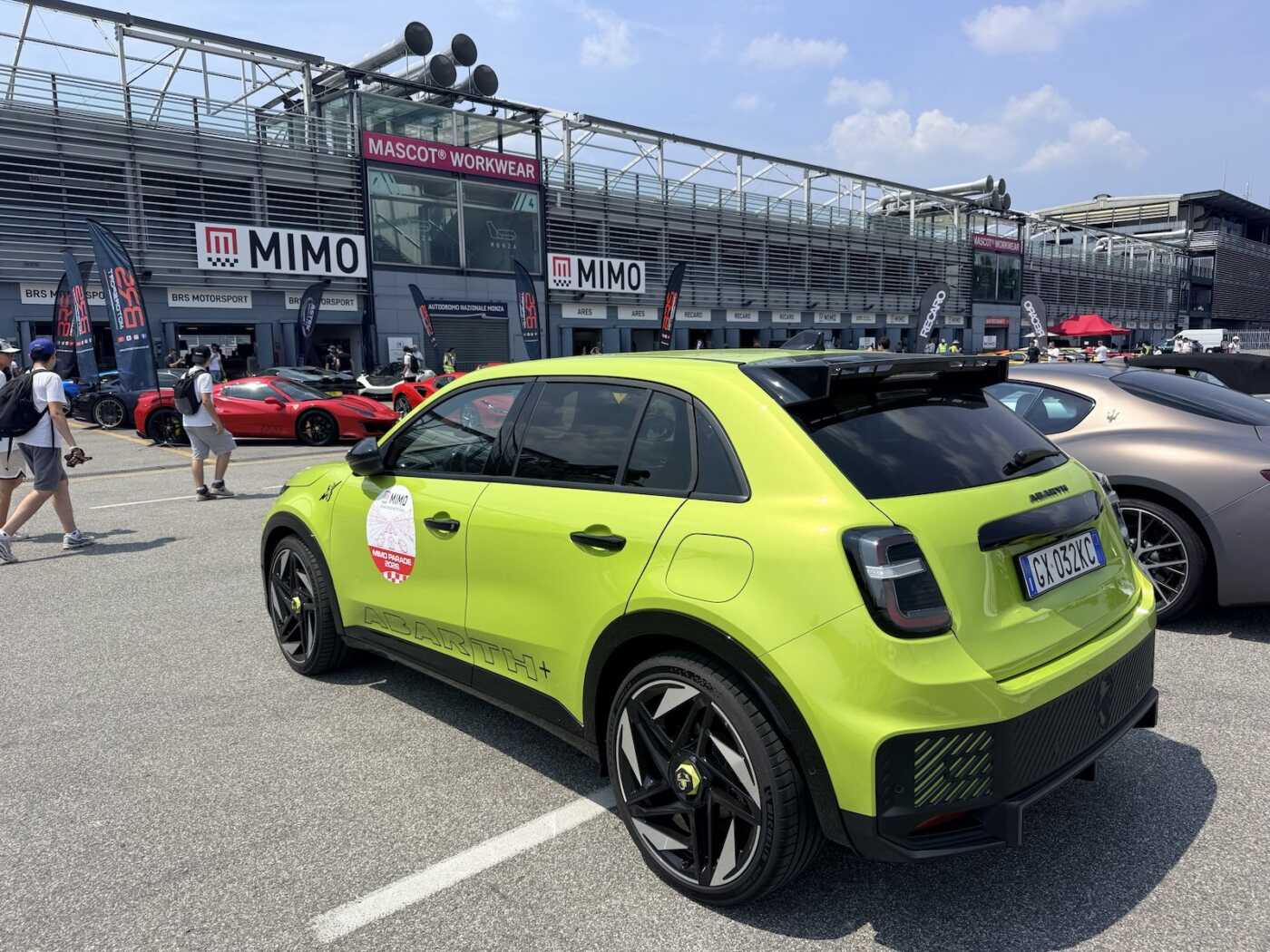 Tre quarti posteriore dell'Abarth 600e Scorpionissima verde acido nel piazzale davanti ai box di MIMO 2026, con le Ferrari sullo sfondo